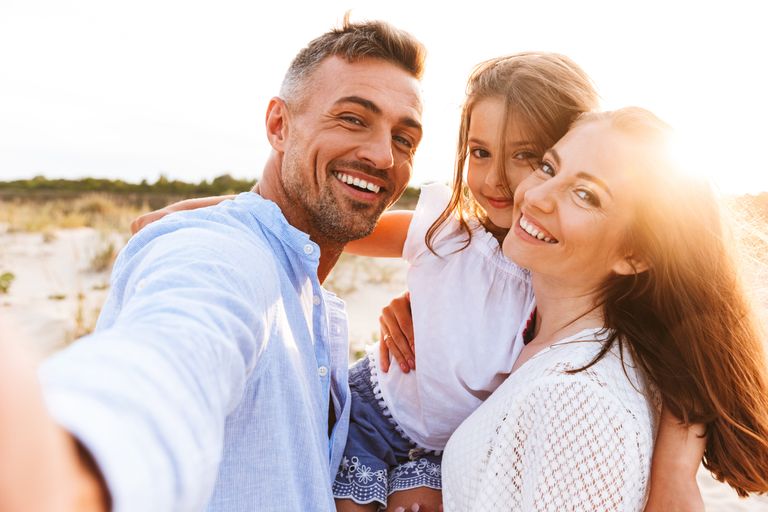 Une famille de 3 personnes, deux adultes et une fille, en selfie avec un fond de plage au coucher du soleil