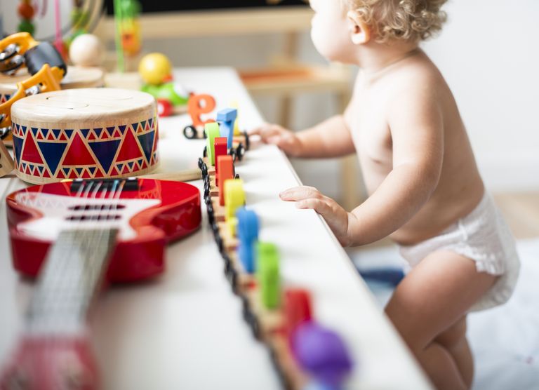 Bébé en couche debout devant une rebord avec des jouets en bois : petit train, tabourin, guitare